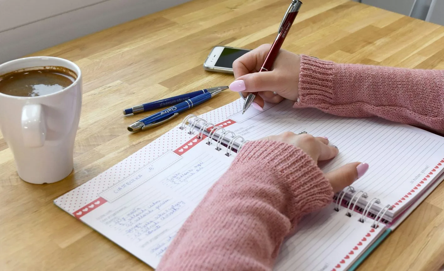 E-learning tanfolyamok – rózsaszín pulóveres kéz jegyzetel a határidőnaplóba, mellette kávé